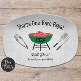 One Rare Papa 10x14 inch grilling platter with gray background and green grill in the center with a rare steak on the grates. BBQ tongs, spatula and fork on the left and right of the grill. Well Done with Love and the names at the bottom