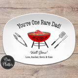 One Rare Dad grilling platter with white background and red grill in the center with a rare steak on the grates. BBQ tools tongs, spatula and fork on the left and right of the grill. Well Done with Love and the names at the bottom