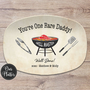 One Rare Daddy 10x14 inch grilling platter with tan background and black grill in the center with a rare steak on the grates. BBQ tongs, spatula and fork on the left and right of the grill. Well Done with Love and the names at the bottom