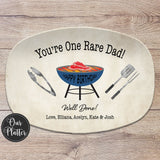 One Rare Daddy 10x14 inch grilling platter with tan background and blue grill in the center with a rare steak on the grates. BBQ tongs, spatula and fork on the left and right of the grill. Well Done with Love and the names at the bottom