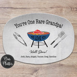 One Rare Papa 10x14 inch grilling platter with blue background and green grill in the center with a rare steak on the grates. BBQ tongs, spatula and fork on the left and right of the grill. Well Done with Love and the names at the bottom